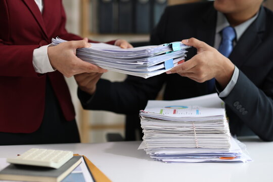 Businessman sitting at his desk, receiving a large stack of documents from his assistant in a busy office, managing a heavy workload.