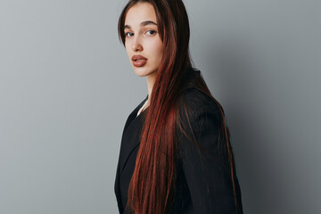 Naklejka premium Young woman with long straight hair wearing black blazer posing and looking at camera with neutral expression on gray background in studio portrait. Female model with natural makeup.