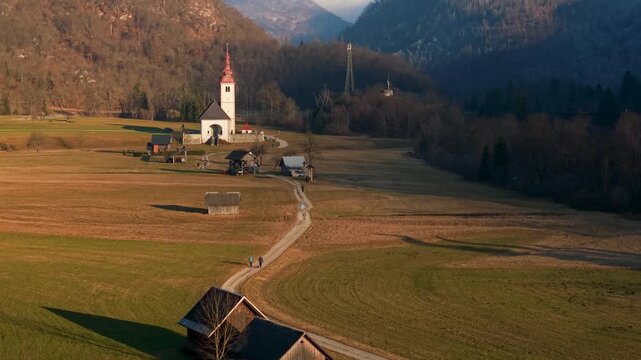 Minimalist alpine road flight towards church with red chapel traditional kozolec hayracks in Bitnje village Bohinj valley Slovenia Julian Alps sunset travel destination dji air 3s  4k