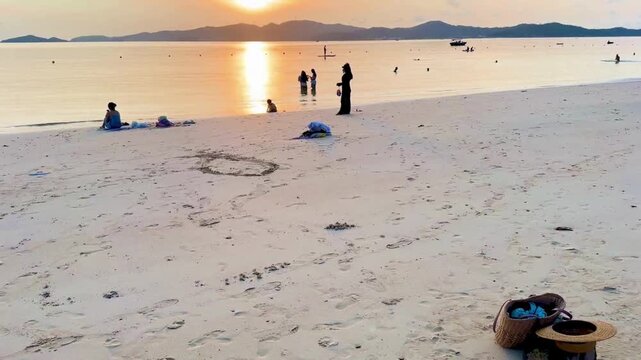 Many pairs of sandals and personal items on the sand while tourists are swimming at sunrise. The video starts with colorful slippers on the beach then expands to a wide view of people swimming