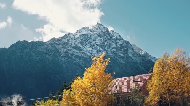 Cinematic view of a snow-capped mountain peak towering over autumn-colored trees and a traditional house in North Ossetia.