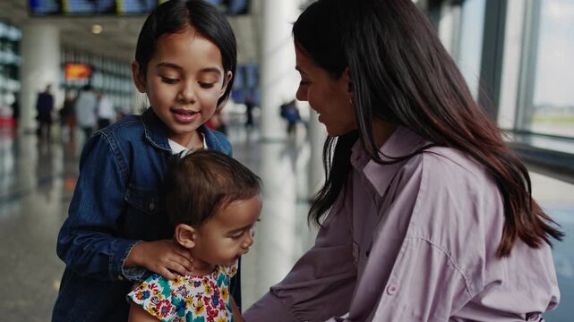 Mother and children at airport window sharing tender goodbye, mother comforts toddler and infant while waiting by glass terminal, luggage and passengers blurred in background, sibling support and warm
