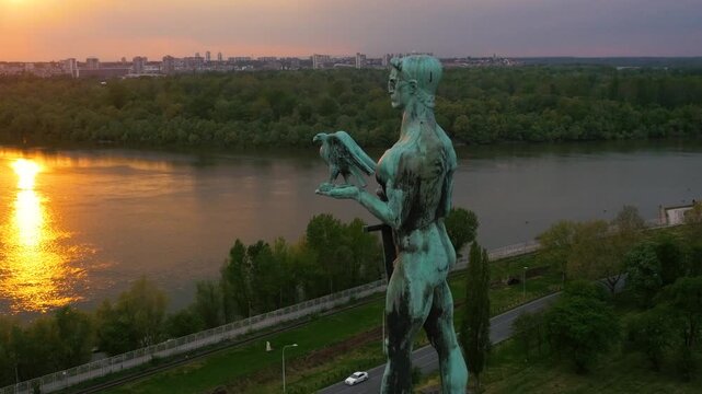 Drone shot of the Victor monument (Pobednik) in Belgrade at sunset. The statue stands over the Sava and Danube confluence, with a glowing golden sky and city skyline in the background.