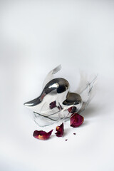 Conceptual still life of a metallic heart among broken glass shards and wilted rose buds on a white background. Symbol of heartbreak, loss and fragile emotions.