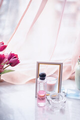Soft feminine still life with perfume bottles, empty photo frame and candles on a light surface. Flowing delicate fabric and pink tulips create a romantic pastel atmosphere with copy space.
