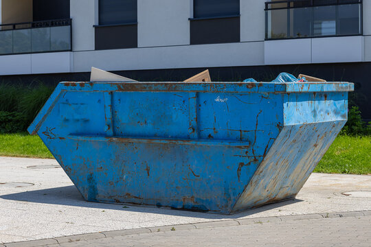 Large blue metal construction dumpster placed near modern residential building on sunny day, industrial waste container for renovation debris and urban cleanup concept