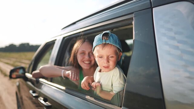 Mother and son look from car window during road trip. Happy family travels on countryside road. Boy with cap looks outside vehicle. Summer vacation journey. Family enjoys road trip together.