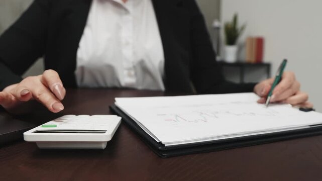 Businesswoman doing calculations on calculator while taking notes on document at her desk in modern office space.