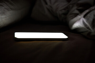Glowing smartphone on bed with dim lighting, illuminating darkness and creating eerie ambience. © elenavolf