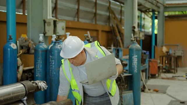 Professional senior male engineer in hard hat using laptop while inspecting heavy machinery and argon gas cylinders in metal factory. Industrial maintenance and quality control concept.