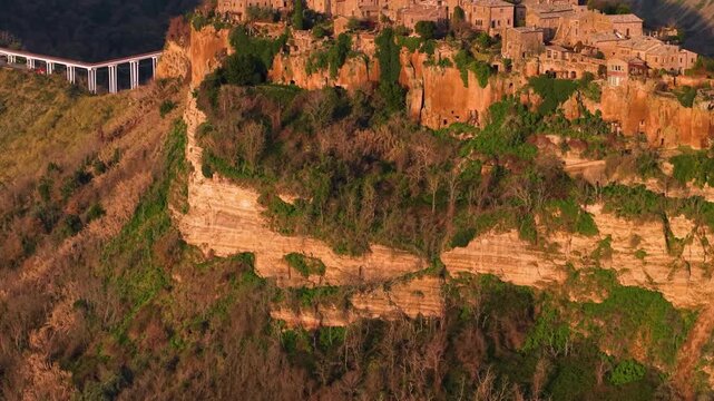 Aerial side reveal of Civita di Bagnoregio at sunrise. Drone view of bridge and steep tuff cliffs in orange morning light, medieval hilltop village Lazio Italy. 4K 10-bit HDR cinematic video.