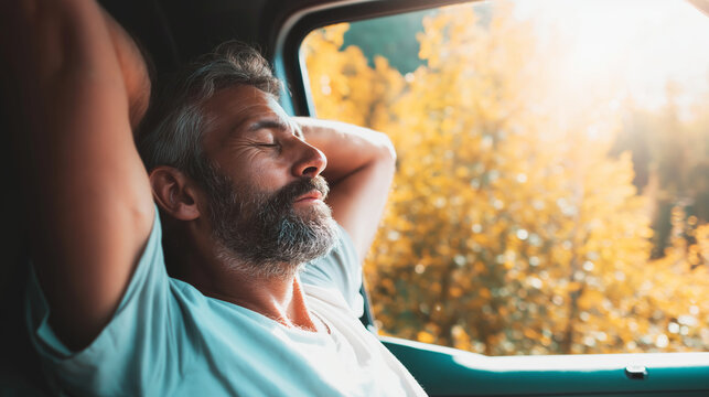 Mature man with closed eyes relaxing inside car, arms behind head, feeling warmth of sunshine and autumn air
