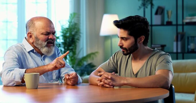 Indian senior father advising young son during serious life discussion while sitting at table in modern living room at home, experienced parent guiding student child about responsibility, future and v