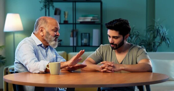 Indian senior father advising young son during serious life discussion while sitting at table in modern living room at home, experienced parent guiding student child about responsibility, future and v
