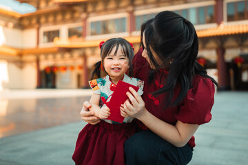 Happy Asian mother and little daughter holding red envelopes or Ang Pao during Chinese New Year. Family celebrating Lunar New Year together at Chinese temple. Cute child and mom in traditional costume