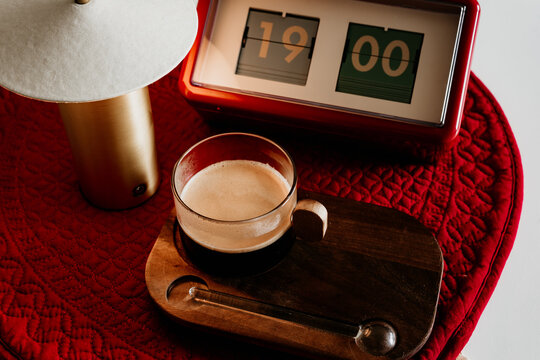 Coffee in a glass cup on a wooden tray with a lamp and a clock showing 19:00 at a cozy indoor setting in the evening