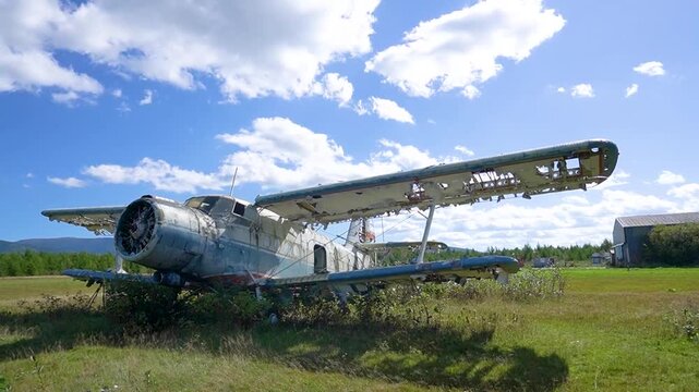 An abandoned agricultural biplane with a radial engine on the territory of an old airfield. The plane is standing in tall grass, the upper wing is partially destroyed, the skin is damaged.