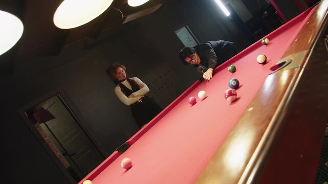 Tilt and dolly shot of pair of youthful caucasian man and woman challenge each other in lively pool game under overhead lamps. Putting a ball into a hole in nightlife billiards club with red table