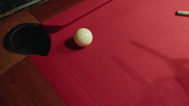 Top view of male player that successfully guides cue ball towards pocket during friendly billiards match. Close up of hand and white ball on red felt table. Casual social gathering in evening club