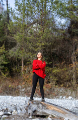 Obraz premium Confident woman in a red sweater stands on a log by a riverbank, surrounded by trees and natural scenery, embodying strength and empowerment in a serene outdoor setting