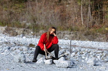 Naklejka premium Young woman in a red sweater sitting on rocky riverbank, thoughtfully resting her chin on hand, surrounded by nature, reflecting on life and tranquility