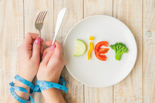 Hands tied with blue measuring tape holding fork and knife over vegetable word diet on plate symbolizing restrictive dieting and body image pressure


