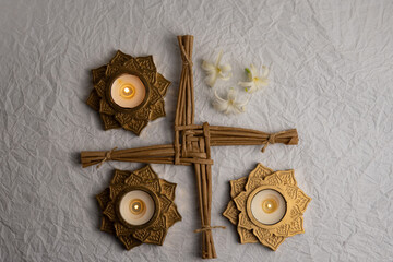 A top view Imbolc altar with a woven Brigid’s cross, golden candle holders, and scattered white flower petals is arranged on a textured light background to symbolize Saint Brigid’s Day.