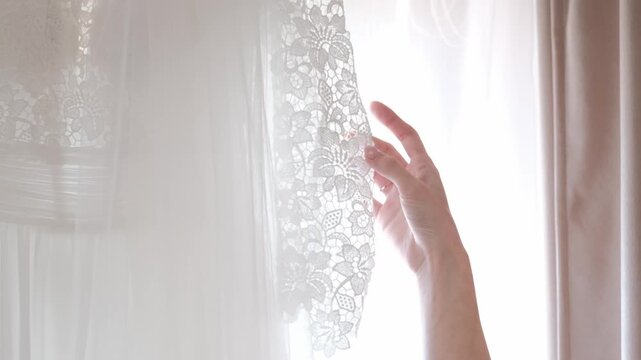 Close-up of a white bridal gown featuring intricate floral lace and sheer tulle fabric hanging near a window.