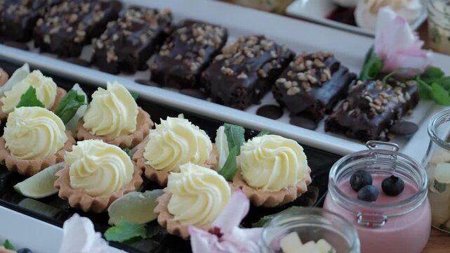 Rows of creamy custard tarts with lime wedges and chocolate brownies topped with nuts on a dessert platter.