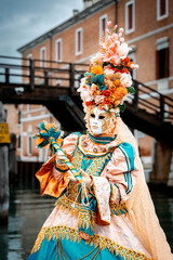 Naklejka premium Venice, Italy - February 09, 2026: Colorful masked figure in elaborate costume with floral headdress poses near a canal and bridge during a vibrant carnival celebration