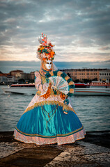 Naklejka premium Venice, Italy - February 09, 2026: Colorful masked performer in elaborate costume holding a fan by the water during a vibrant carnival celebration at sunset