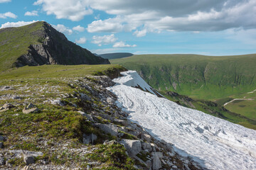 Scenic sunlit view along grassy ridge with white snow cornice to green hill top with rocky sheer crags in bright sun under lush clouds in blue sky. Vivid alpine scenery in high mountains in sunny day. © Daniil
