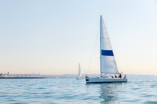 Sailboat With White Sails and Blue Stripe Sailing on Calm Sea Near Coastal City at Sunrise