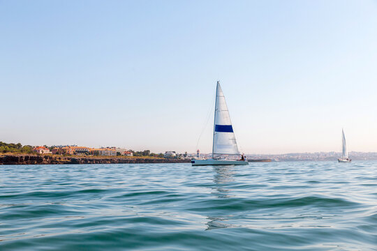 Sailboat With White Sails and Blue Stripe Sailing on Calm Sea Near Coastal City at Sunrise