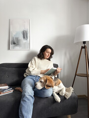 Cozy home education young woman reading a book on a sofa with her beagle dog at home. © Iryna