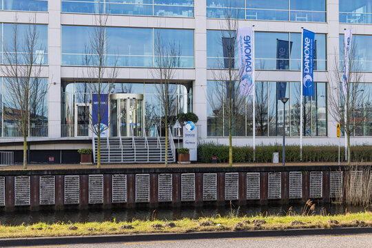 The entrance of the Danone Nutricia headquarters. Features corporate flags, revolving doors, and a canal in the foreground. Hoofddorp, Netherlands. 8 February 2026.