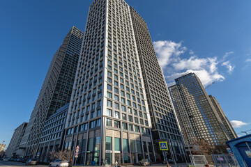Obraz premium Tall buildings reach for the sky in a busy city area. The scene captures a clear day with blue skies and a few clouds above the modern structures. People walk on the street below.