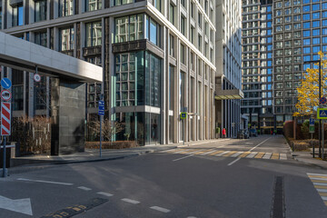 Obraz premium People walk along the street near tall buildings. Cars are parked on the side. The sun shines on the glass facades. Trees provide some color to the scene.