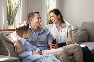 Obraz premium Laughing Caucasian parents and child sitting on couch in living room, holding smartphone, lead positive conversation, discussing new mobile application. Family enjoy time together on leisure at home
