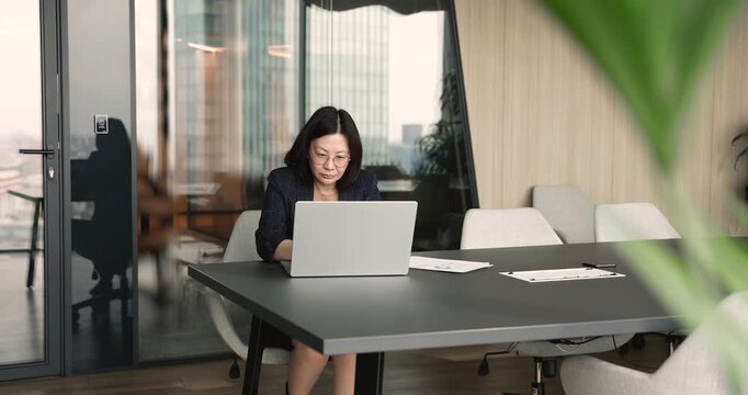 Asian businesswoman or executive sits at table, working on laptop, focused in business tasks, reviewing report or digital documents. Data analysis, planning, consulting, corporate workflow in office