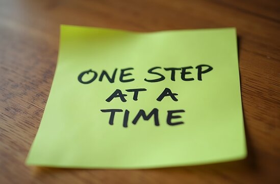 green sticky note placed on a wooden desk with the phrase &lsquo;ONE STEP AT A TIME&rsquo; written in neat black handwriting