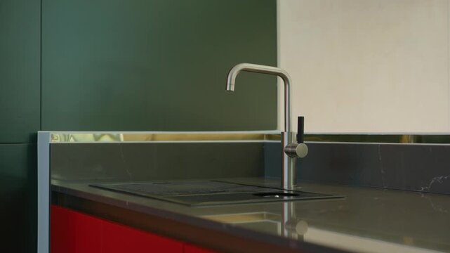 The kitchen design is modern and minimal, dark countertop holds sleek stainless steel faucet with curved spout and lever handle, backsplash combines green and beige panels