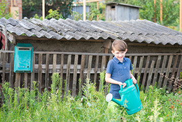 Little boy holding green watering can in a traditional Russian village. Cute 8 year old child helping with gardening in countryside backyard near old wooden fence and rustic barn. © Ruslan Russland