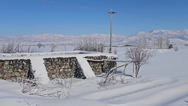 Snowy rural landscape with an agricultural water tank, a long stone wall, scattered trees, and distant mountains under a clear blue sky creating a calm winter scene.  
📍Marivan, Kurdistan 