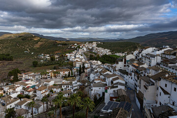 Paisaje urbano de Izn&aacute;jar, uno de los pueblos m&aacute;s bellos de Espa&ntilde;a, con sus casas blancas tradicionales sobre una colina rodeada de extensos olivares bajo un cielo nublado en Andaluc&iacute;a