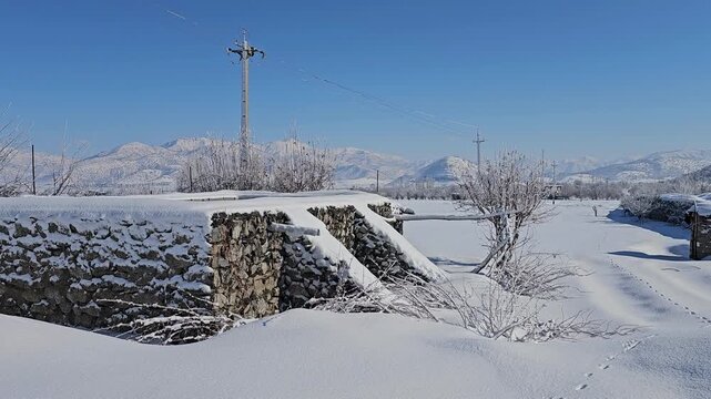 Snowy rural landscape with an agricultural water tank, a long stone wall, scattered trees, and distant mountains under a clear blue sky creating a calm winter scene.  
📍Marivan, Kurdistan 