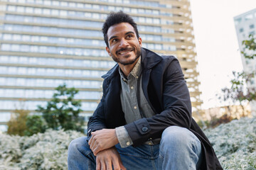 Fototapeta premium Young adult Latin American man smiling while seated outdoors. Relaxed professional posing confidently in modern city park. Lifestyle and wellbeing concept.