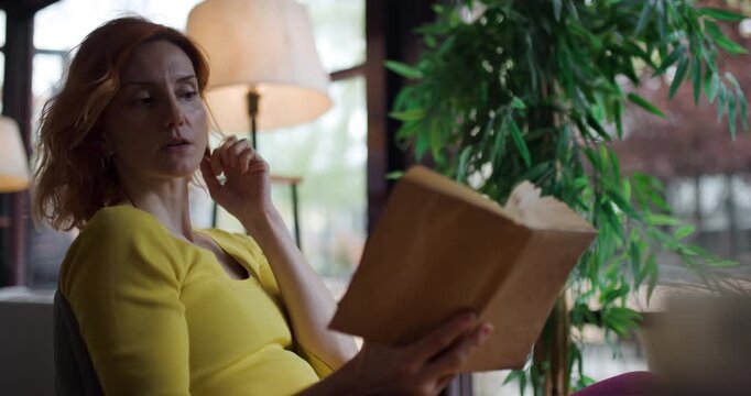 Woman reading a book by window during the afternoon at cafe, wearing a yellow top while relaxing near green plants in the background