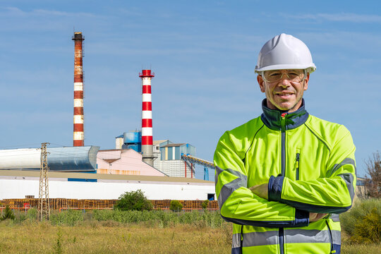 Industrial worker wearing PPE with arms crossed in front of a factory chimneys