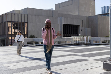 Adult woman walking on terrace in pink hoodie, talking on phone, carrying backpack, crossbody bag